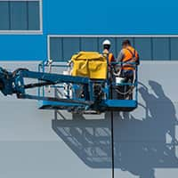 A warehouse worker in a safety vest and helmet stands on a ladder while organizing boxes on a high shelf.