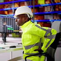 A man in a hard hat, sitting in a desk chair holding his back