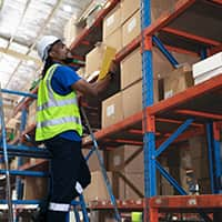 A warehouse worker in a safety vest and helmet stands on a ladder while organizing boxes on a high shelf.