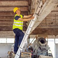 A worker in a safety vest and helmet stands on a ladder on a construction site.