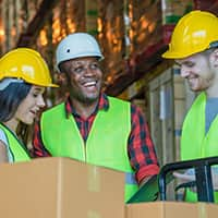 A warehouse worker in a safety vest and helmet stands on a ladder while organizing boxes on a high shelf.