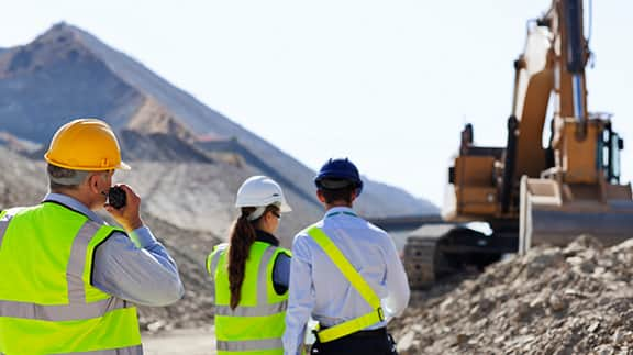 Three construction workers in safety vests and hard hats stand near a construction site, overseeing ongoing work activities.