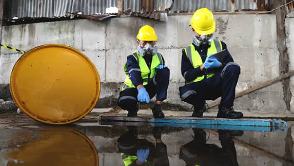 Two men wearing yellow safety gear are kneeling next to a puddle, assessing the situation for potential hazards.