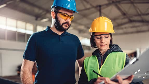 A man and woman in hard hats and safety glasses examine a clipboard, discussing safety protocols on a construction site.