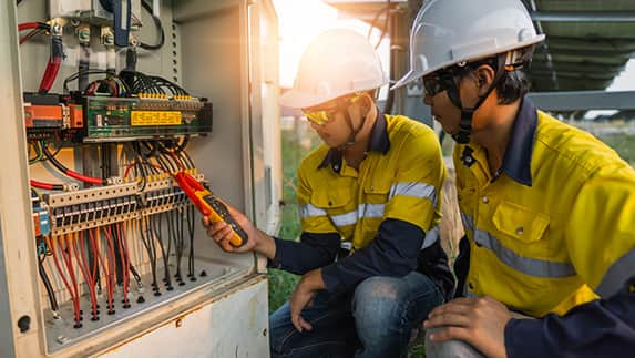 Two workers in yellow shirts and hard hats are engaged in maintenance on an electrical panel.