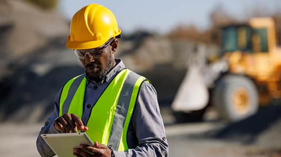 A construction worker in a hard hat and safety vest is using a tablet on a job site.