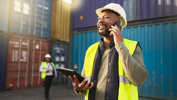 A man wearing a hard hat and safety vest is engaged in a conversation on his cell phone.