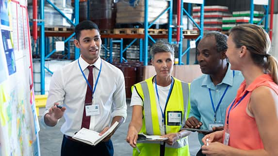 A group of individuals in a warehouse examining a map, engaged in discussion and planning their next steps.