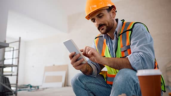 A man wearing a hard hat and safety vest is focused on using a tablet at a construction site.