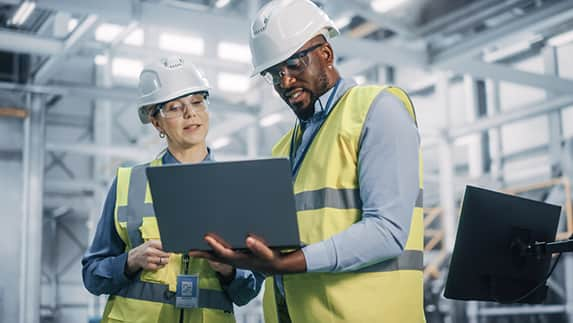 Two workers in hard hats and safety vests examine a laptop together on a construction site.